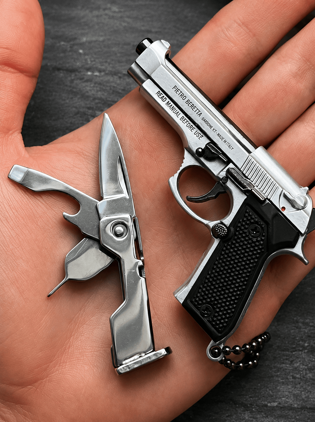 Metallic pocket knife and gun-shaped keychain on a hand against a dark background