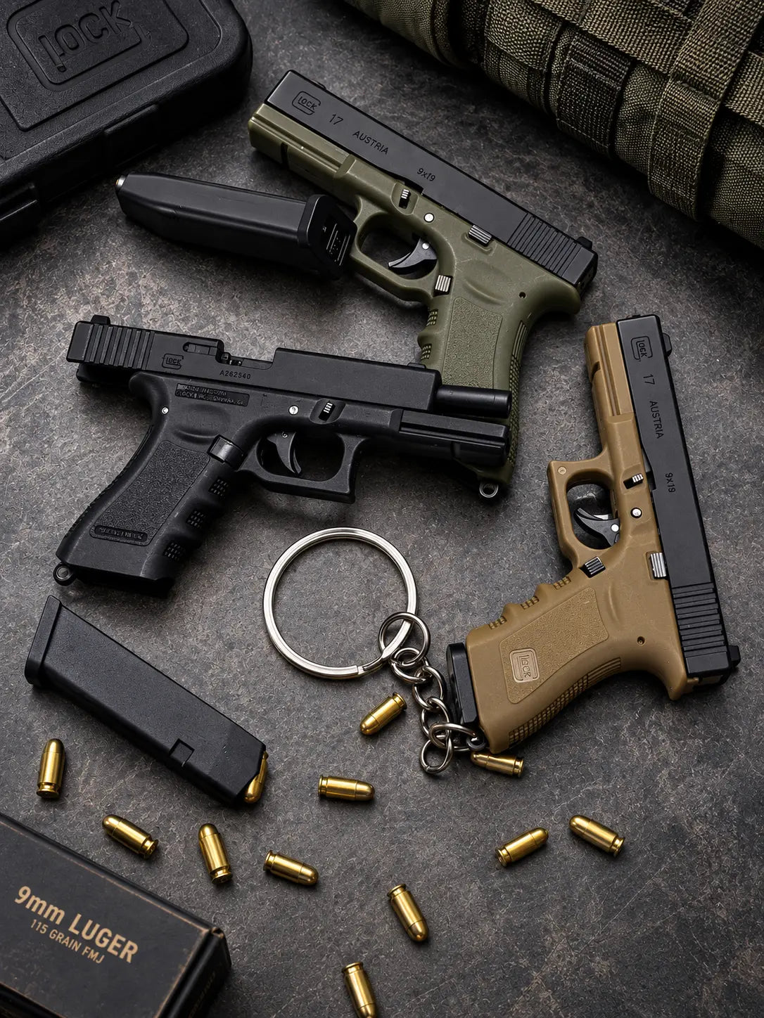 Three Die-Cast keychains s in black, olive green, and tan are arranged on a dark textured surface with bullet casings, magazines, a keychain, and a box.