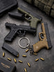 Three Die-Cast keychains s in black, olive green, and tan are arranged on a dark textured surface with bullet casings, magazines, a keychain, and a box.