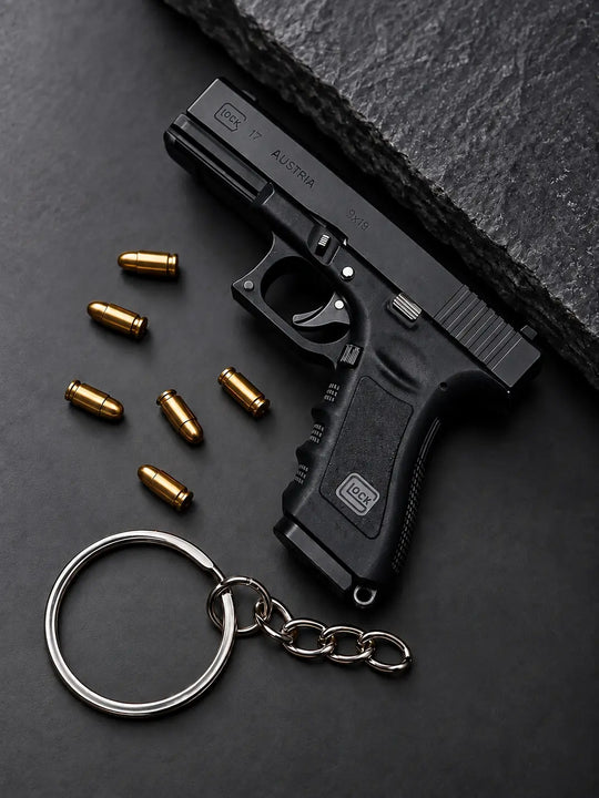A black Glock Die-Cast -shaped keychain with a silver split ring and chain, displayed on a dark textured surface next to several gold-colored bullets. The Glock logo is visible on the grip and slide.