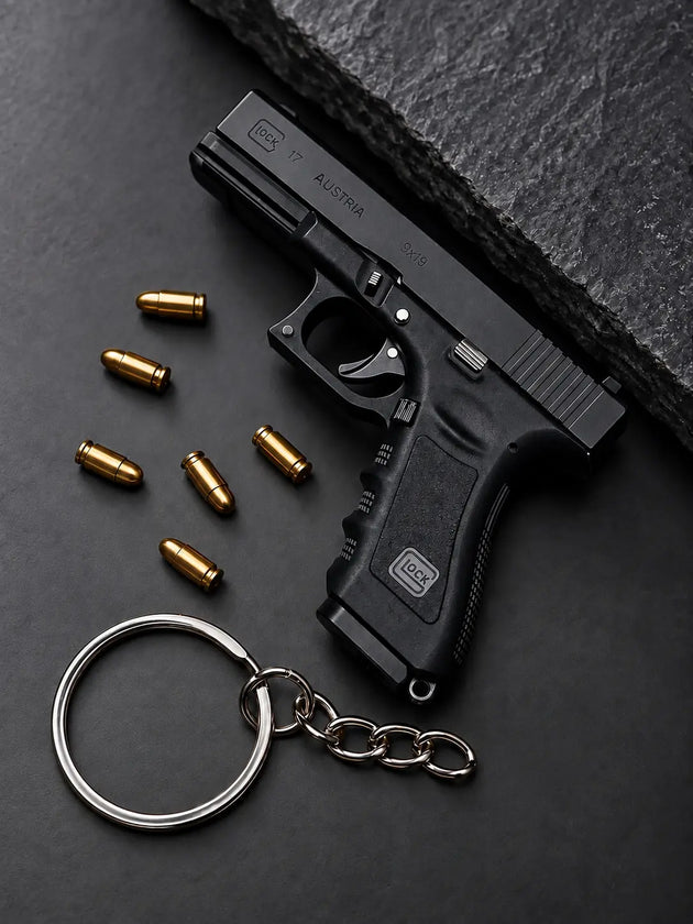 A black Glock Die-Cast -shaped keychain with a silver split ring and chain, displayed on a dark textured surface next to several gold-colored bullets. The Glock logo is visible on the grip and slide.