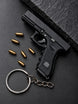 A black Glock Die-Cast -shaped keychain with a silver split ring and chain, displayed on a dark textured surface next to several gold-colored bullets. The Glock logo is visible on the grip and slide.