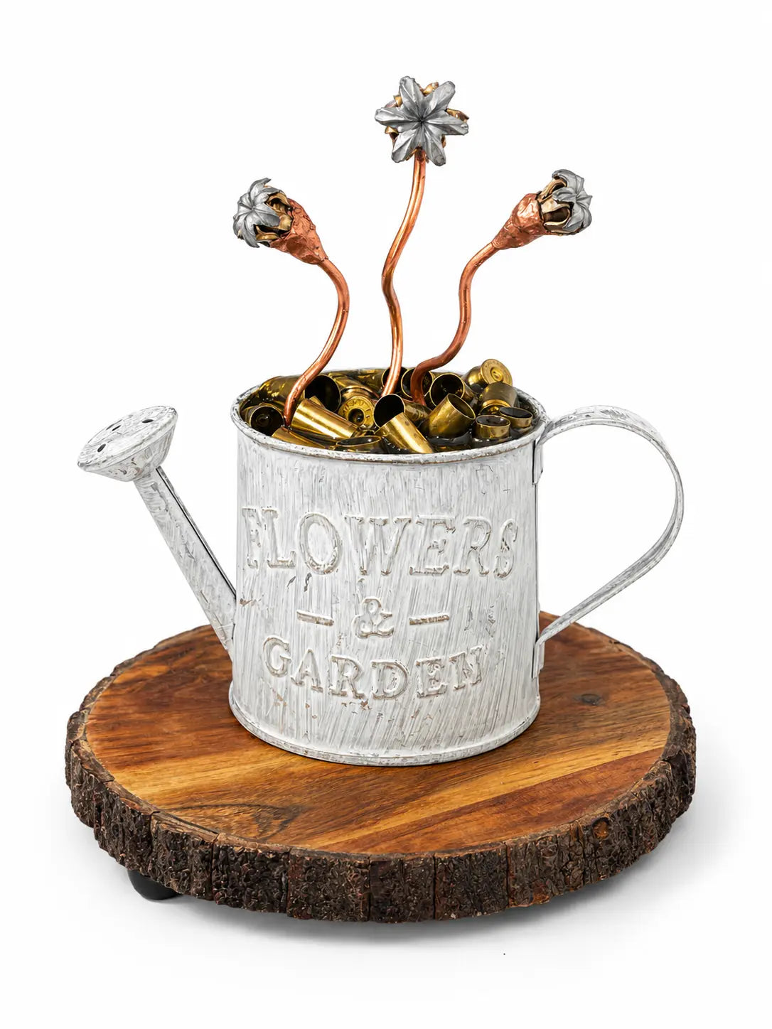 A decorative sculpture of a galvanized metal watering can engraved with 'FLOWERS & GARDEN,' filled with small metallic bells and topped with three copper-colored stems ending in flower buds, displayed on a round wood slice base against a white background.