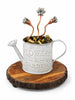 A decorative sculpture of a galvanized metal watering can engraved with 'FLOWERS & GARDEN,' filled with small metallic bells and topped with three copper-colored stems ending in flower buds, displayed on a round wood slice base against a white background.