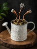 A decorative metal watering can labeled “FLOWERS & GARDEN,” filled with small gold and bronze flower buds and topped with three copper stems ending in flower-shaped tips, displayed on a round wooden slice against a dark background.