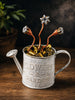 A decorative galvanized metal watering can labeled 'FLOWERS & GARDEN' filled with miniature flower bulbs and copper-and-silver flower stems, set on a wooden surface with a dark blurred background.