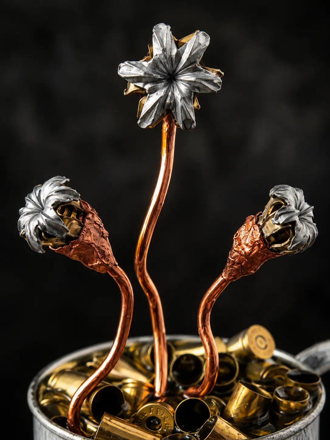 A decorative sculpture of three metallic flower-like forms rising from a metal container filled with brass bullet casings, photographed against a dark black background.