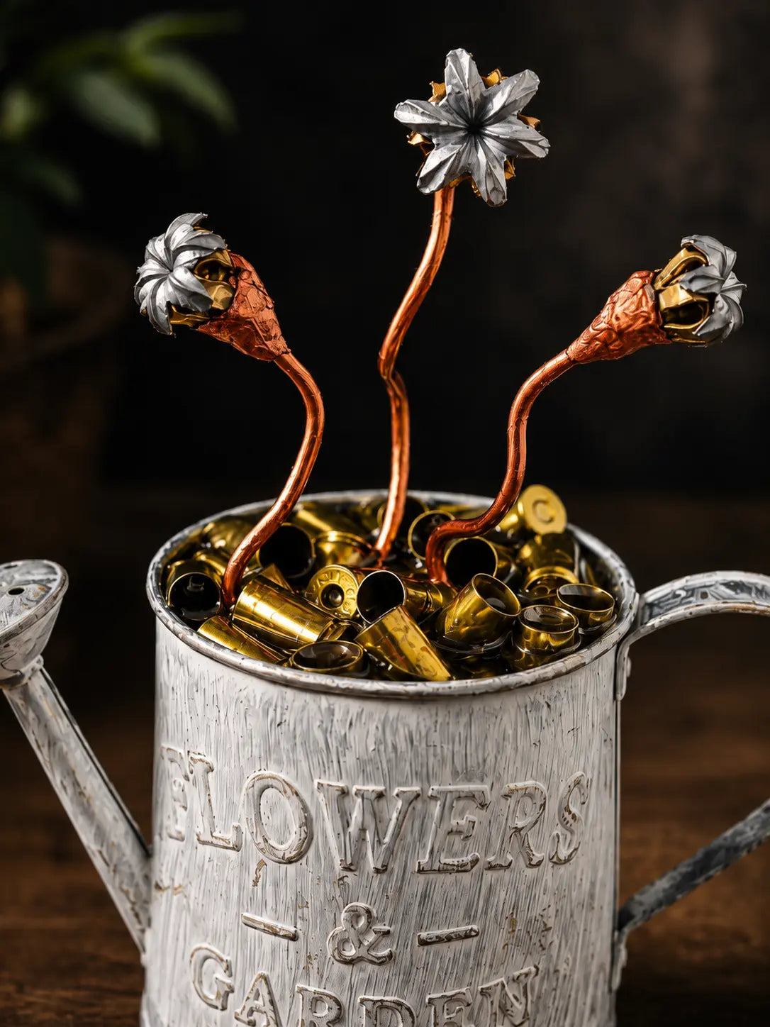 A rustic silver metal watering can engraved with 'FLOWERS & GARDEN,' filled with brass bullet casings and topped with three copper and silver flower-shaped ornaments.