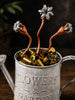 A rustic silver metal watering can engraved with 'FLOWERS & GARDEN,' filled with brass bullet casings and topped with three copper and silver flower-shaped ornaments.