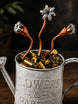 A rustic silver metal watering can engraved with 'FLOWERS & GARDEN,' filled with brass bullet casings and topped with three copper and silver flower-shaped ornaments.