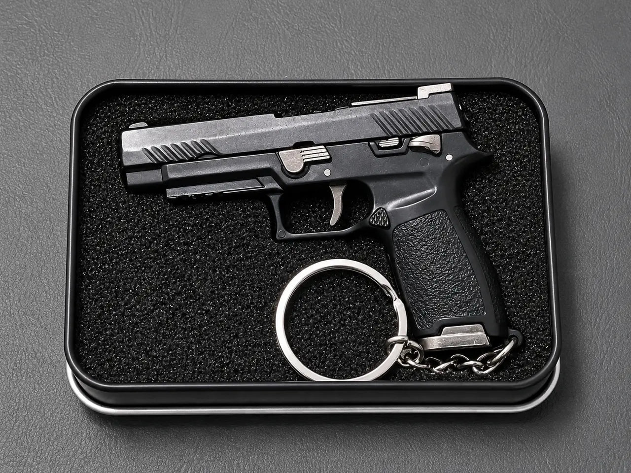 A black semi-automatic pistol keychain displayed inside a black rectangular presentation box with a textured foam insert, set against a dark gray background.