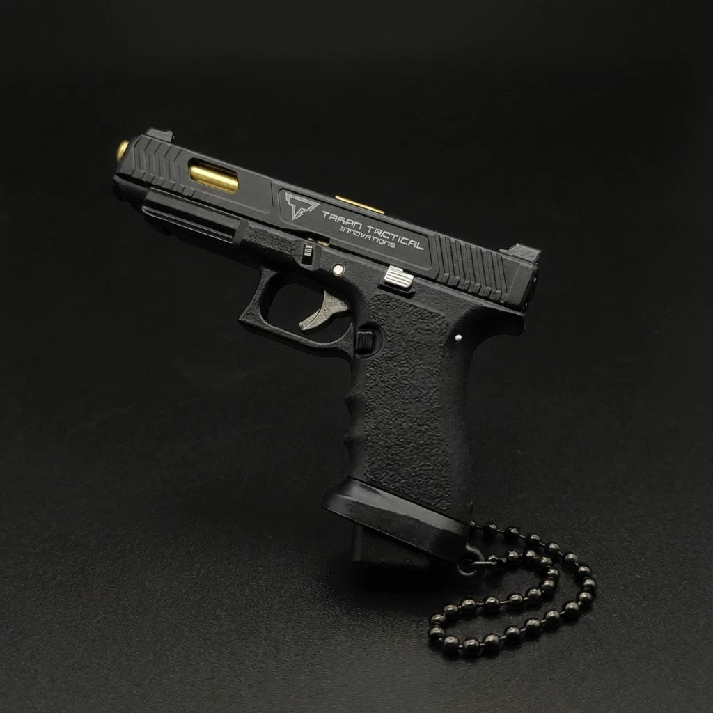 A black semi-automatic pistol with gold accents and a black beaded chain, photographed on a dark black background. The slide features the 'Tactical Tactical' logo.