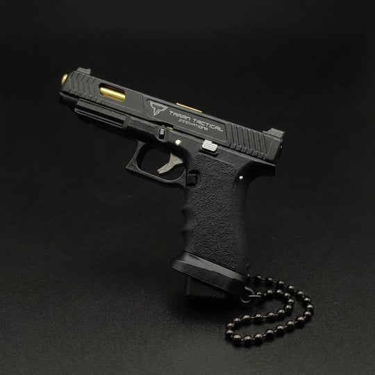 A black semi-automatic pistol with gold accents and a black beaded chain, photographed on a dark black background. The slide features the 'Tactical Tactical' logo.
