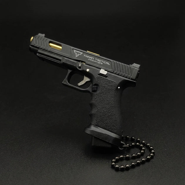 A black semi-automatic pistol with gold accents and a black beaded chain, photographed on a dark black background. The slide features the 'Tactical Tactical' logo.