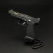 A black semi-automatic pistol with gold accents and a black beaded chain, photographed on a dark black background. The slide features the 'Tactical Tactical' logo.
